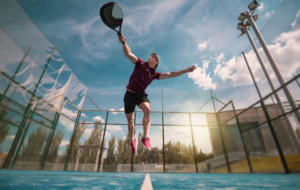 Hráč padelu skákající ve vzduchu při úderu na míček na venkovním kurtu, s modrou oblohou a slunečním světlem v pozadí.