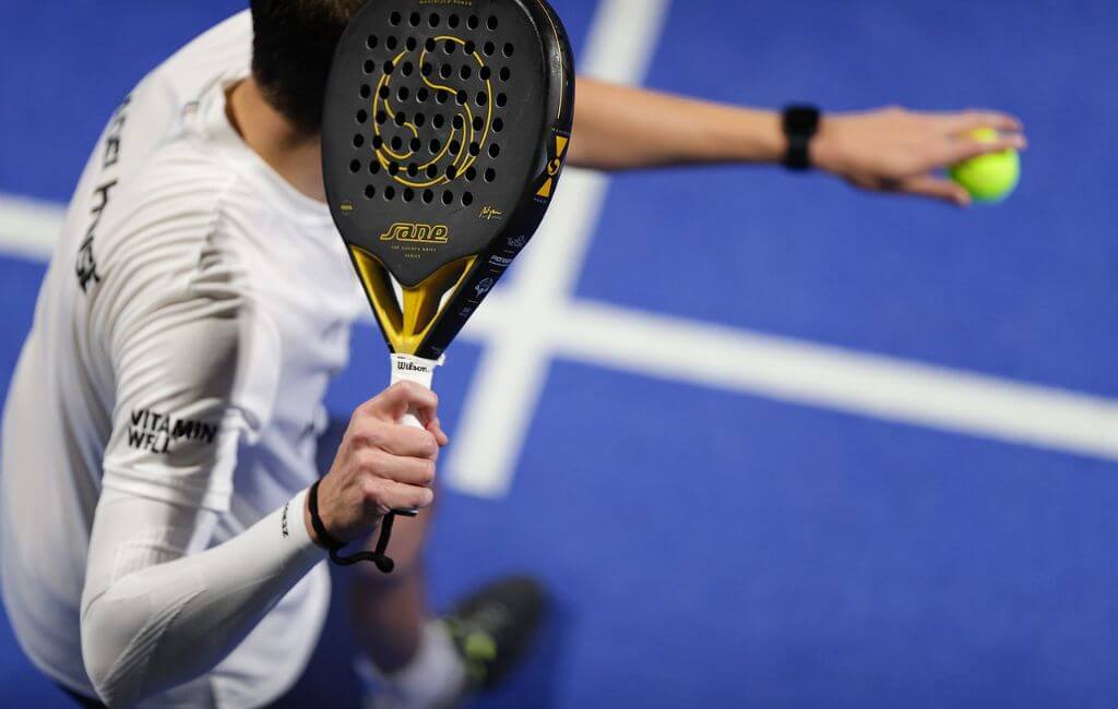 Detailní záběr hráče padelu, který drží raketu a míček, připravený ke hře na modrém kurtu.